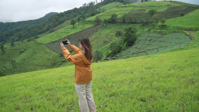 年轻美丽的亚洲女子徒步旅行者黄夹克站在山顶用智能手机拍摄美丽的自然森林山和草山的慢动作。科技与度假概念。视频下载