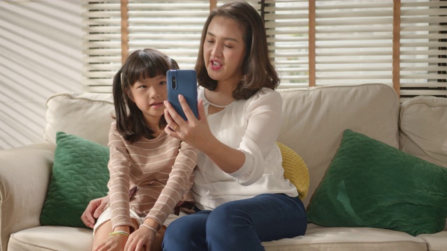 快乐的亚洲家庭，妈妈和小女儿用智能手机视频聊天或自拍，坐在沙发上，在家里的客厅里享受在线交流。视频素材
