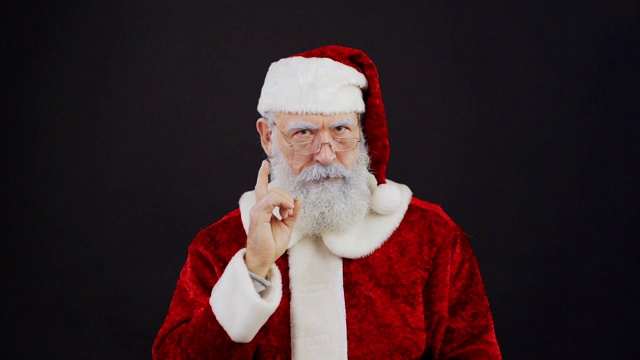 腰部以上的圣诞老人肖像在传统的红色服装威胁食指和调整眼镜在慢动作站在黑色演播室背景视频下载