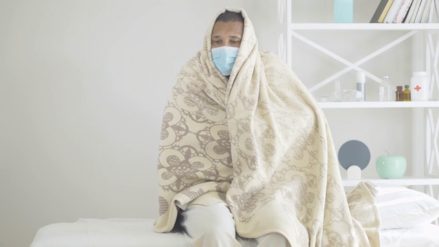 生病的悲伤的人戴着面具，裹着毯子坐在医院病房里。患Covid-19热的非裔美国男性患者画像。冠状病毒病的症状。视频素材