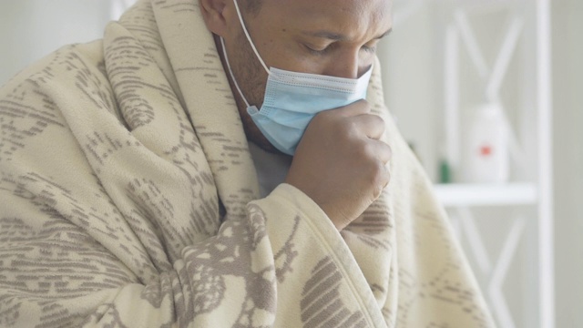 特写咳嗽的非裔美国人在面罩包裹在毯子。伴有Covid-19病毒病症状、疲惫不堪的男性患者画像。冠状病毒的爆发。视频素材