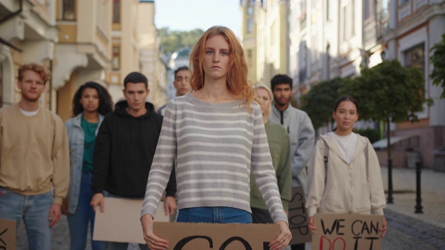 一名红发女子站在前面，她展示了抗议种族主义的海报，背景是学生的抗议视频素材