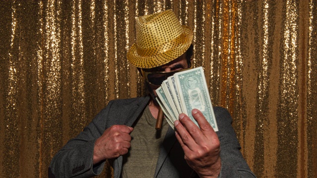 一个留着胡子的男人戴着帽子，抽着雪茄，拿着一堆美钞在照相亭里摆姿势视频素材