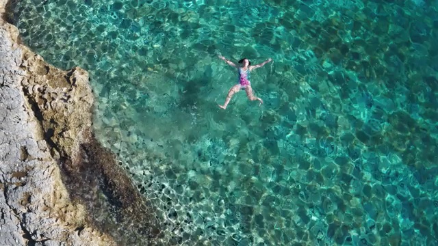 年轻的女性漂浮在背上，放松在温暖的碧绿的亚得里亚海海浪与岩石海岸。悠闲的人度假时间概念空中俯瞰4K镜头。视频素材