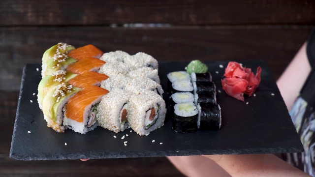 女服务员在黑色的木墙背景上展示一盘寿司，里面有金枪鱼、鲑鱼、黄瓜和鳄梨。视频素材