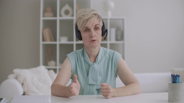 一位老年妇女正用麦克风对着耳机，看着摄像头，坐在家里的桌子上，呼叫中心和视频会议技术视频素材