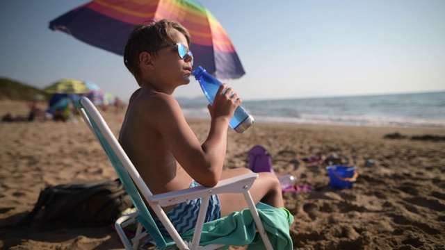 沙滩上的小男孩用可重复使用的水瓶喝水。视频下载