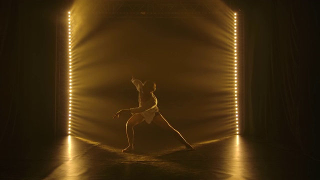 一位身材苗条的年轻芭蕾舞演员正在动情地跳着现代舞。在黑暗的录音室里拍摄，有黄色的灯光和烟雾。轮廓。慢动作视频下载