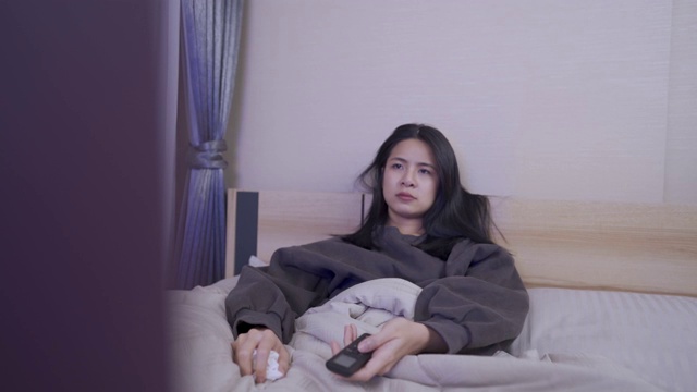 亚洲年轻女子晚上躺在床上的毯子里看电视失眠，生病在卧室，咳嗽在纸巾上，家庭娱乐电视，冠状病毒covid-19视频素材