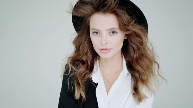 一个时尚的年轻女子在白色背景下的工作室肖像。模特戴着黑色的帽子和衬衫在镜头前摆姿势视频下载