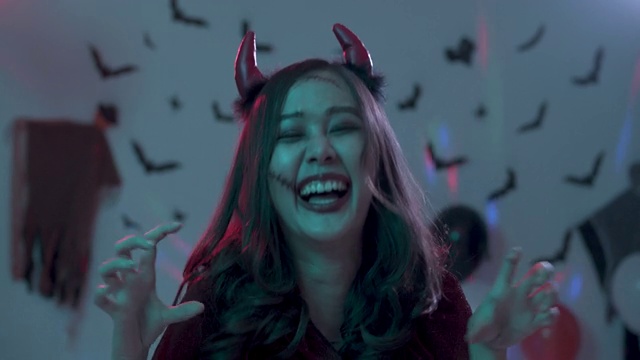 近距离拍摄，迷人的年轻亚洲女人的肖像穿着吸血鬼服装看着镜头微笑，摆姿势在夜晚的万圣节派对在黑暗的背景与彩色的灯光设置。视频购买