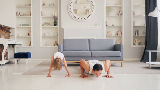 运动时间在家里的客厅，爸爸和他的小可爱的儿子一起做俯卧撑，而小男孩在他的背上。视频素材