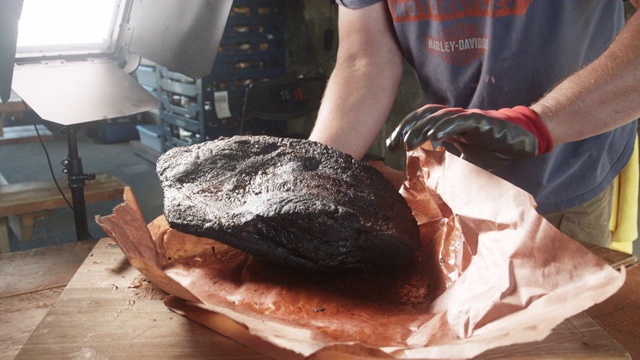 一名男子正在打开一个新鲜的烟熏烤牛胸肉，放在一个简陋的木砧板上，准备切成片视频下载