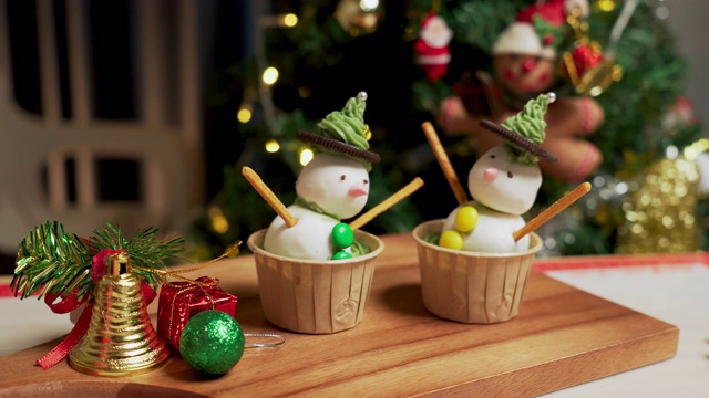 在一个装饰着圣诞树和装饰品的厨房里，两个自制的纸杯蛋糕用奶油色的帽子装饰，用巧克力增添眼睛视频素材