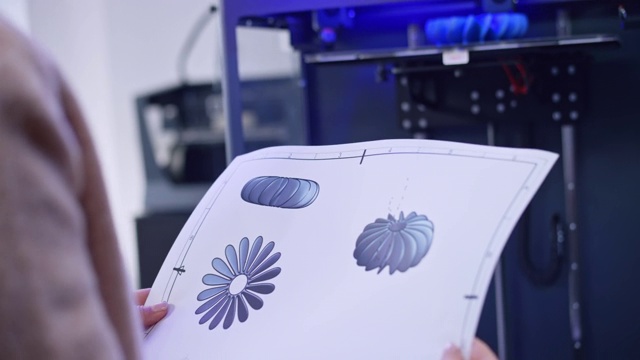 一位女性工程师站在3D打印机旁边，手里拿着一个风扇螺旋桨的打印设计图视频下载