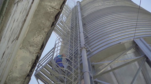 穿着制服和安全帽的人在巨大的水泥仓库行走的广角镜头。巨大的不锈钢塔，白人工人用梯子爬上去。视频下载