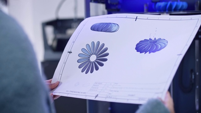 一名女性工程师站在3D打印机旁边，手里拿着一张风扇螺旋桨设计的打印纸视频下载
