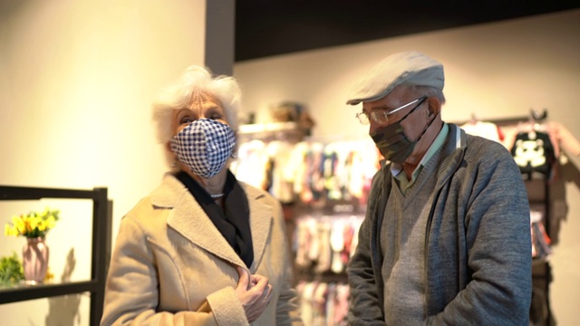 一位戴着口罩的老人在旧货店帮妻子试穿外套视频下载