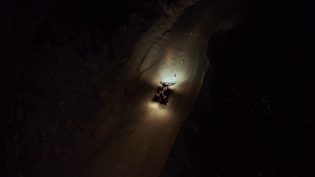 在夜晚的黑暗中，被橙色灯光照亮的雪车沿着白雪皑皑的山丘行驶。视频下载