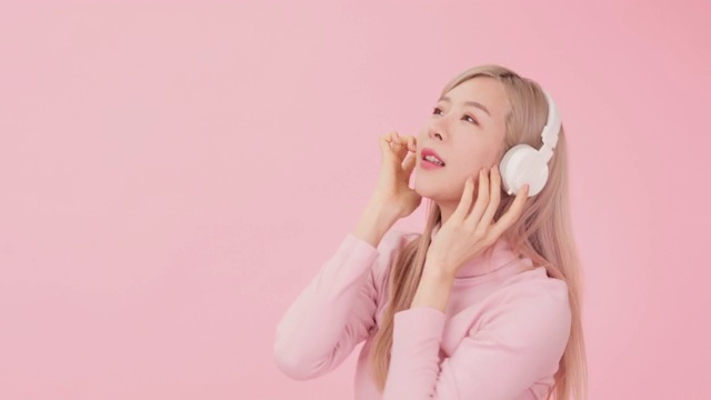韩国女孩的肖像听音乐通过耳机和舞蹈孤立在工作室的粉红色背景。视频素材