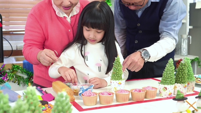 东南亚家庭欢度圣诞时光，一起庆祝圣诞装饰家用装饰品。正面的十几岁的孙女在爷爷和奶奶的帮助下装饰自制的绿树纸杯蛋糕视频下载