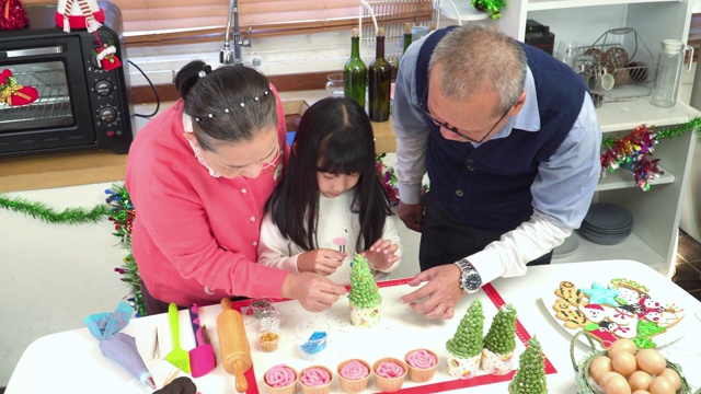 东南亚家庭欢度圣诞时光，一起庆祝圣诞装饰家用装饰品。高视角:十几岁的孙女在爷爷和奶奶的帮助下装饰自制的绿树纸杯蛋糕视频下载