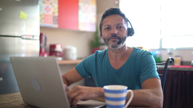 一个成年男子在家工作的肖像，戴着耳机，使用笔记本电脑视频下载