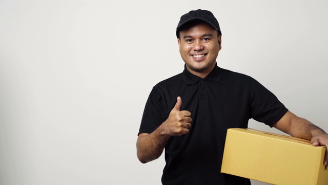 年轻的亚洲快递员在黑色制服拿着包裹纸板指向空白的手指在孤立的白色背景上的文字。4 k决议。视频下载