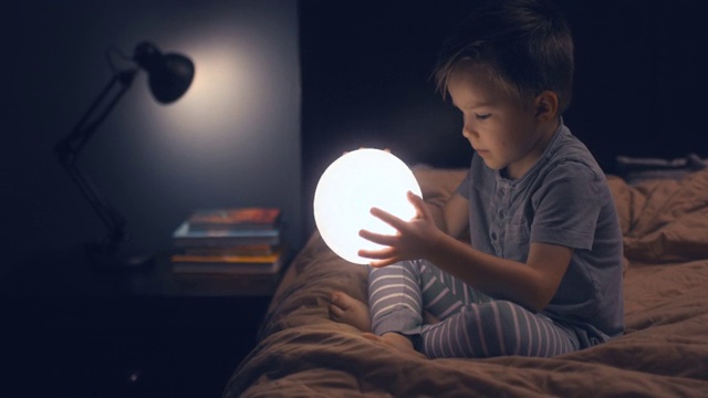 一个小孩坐在床上，手里拿着一轮发光的月亮。视频下载