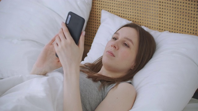 女人在早上坐在卧室靠在柔软的皮革床背。用你的智能手机写信息，使用应用程序来获取信息视频素材