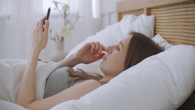 女人在早上坐在卧室靠在柔软的皮革床背。用你的智能手机写信息，使用应用程序来获取信息视频素材
