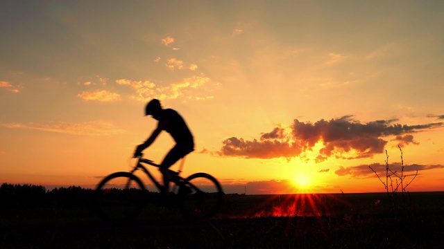 骑自行车的人骑着自行车在夕阳的背景下。一个骑着自行车的人在乡下的剪影。视频下载