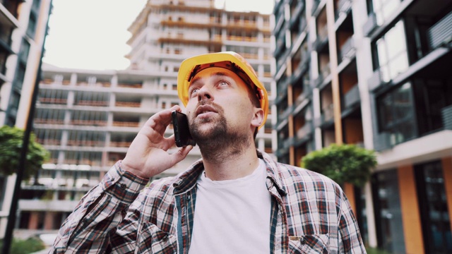 白种男建筑工人橙色安全帽和格子衬衫在建筑工地打电话。建筑的主题。男性的职业。工头通过智能手机控制施工过程。视频素材