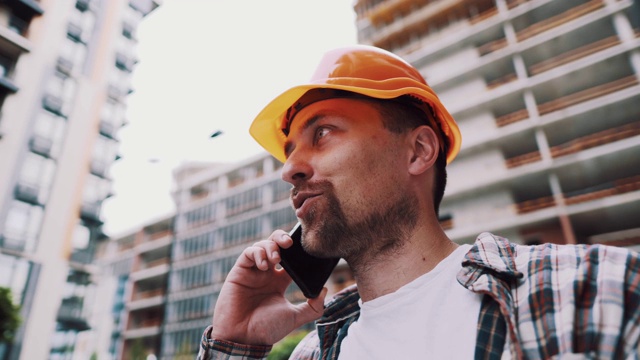 白种男建筑工人橙色安全帽和格子衬衫在建筑工地打电话。建筑的主题。男性的职业。工头通过智能手机控制施工过程。视频素材