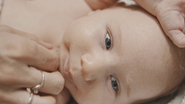 一个蓝眼睛的小婴儿躺在床上，他的妈妈在他的脖子上涂霜视频素材