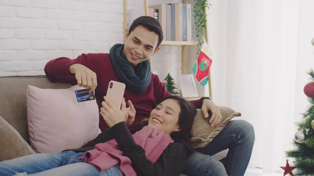 年轻的亚洲情侣情侣一起坐在沙发上。在以圣诞树为背景的房间里，用信用卡和智能手机在网上购物。4 k慢动作。视频素材