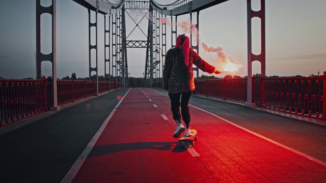 年轻的潮女在非正式的服装是滑板在桥上拿着燃烧的红色信号弹和挥舞它视频购买