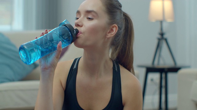 运动女性在训练后在家休息时喝水和使用手机。健康的生活方式。伸展,身体前倾。训练距离。视频下载