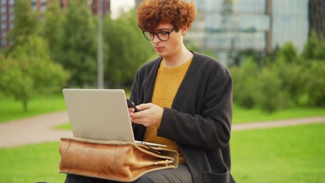 镜头拉近:一个戴眼镜、红卷发的年轻人坐在户外的长椅上，一边用手机发短信，一边思考。打开笔记本电脑放在他腿上的公文包上视频素材