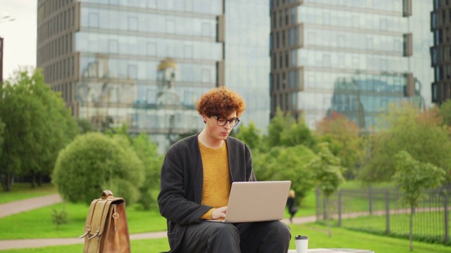 放大镜头，一个红头发的卷发男子坐在户外的长椅上，一边用笔记本电脑工作，一边若有所思地看着别处视频素材