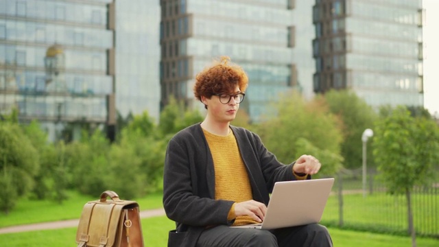 放大镜头，一个戴着眼镜，留着红色卷发的年轻人坐在户外长椅上，用着笔记本电脑，关上电脑，若有所思地看着别处视频素材