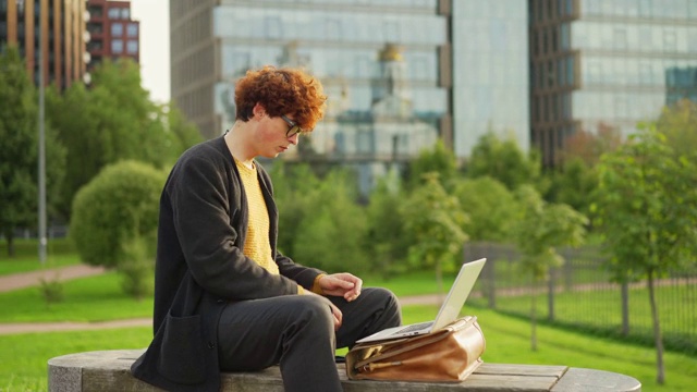 放大侧面镜头，一个留着红卷发的时髦年轻人坐在户外的长椅上，在放在公文包上的笔记本电脑上打字，在手机上浏览信息，喝着咖啡，思考着视频素材