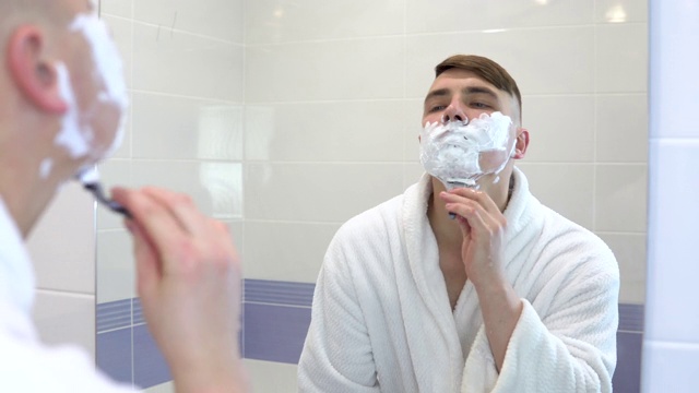 一个年轻人对着镜子刮他的脸毛。一名身穿白大褂、脸上涂着泡沫的男子剃光了头发。透过镜子观察。视频下载
