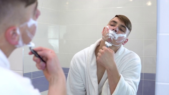 一个年轻人对着镜子刮他的脸毛。一名身穿白大褂、脸上涂着泡沫的男子剃光了头发。透过镜子观察。视频素材