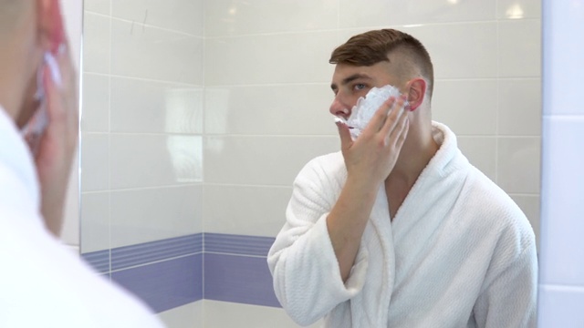 一个年轻人在镜子前涂抹剃须泡沫。一个穿白大褂，脸上有白沫的人。透过镜子观察。视频素材