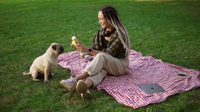 微笑的女孩拍照与可爱的哈巴狗在绿色城市公园拿着相机，坐在格子视频素材