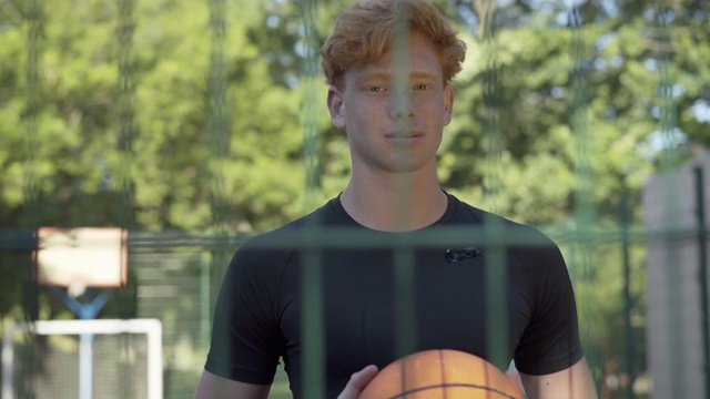年轻红发运动员的肖像站在网状围栏后，看着相机。英俊的白人男子摆姿势与篮球户外在阳光明媚的夏天。体育的概念。视频素材
