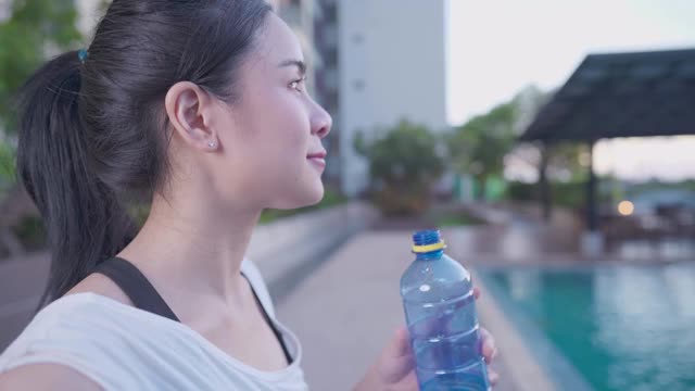 活跃的亚洲女性在运动后从瓶子里喝水，站在游泳池旁，放松下午运动休息，矿泉水，保持水分健康的生活方式健康，侧视图视频素材