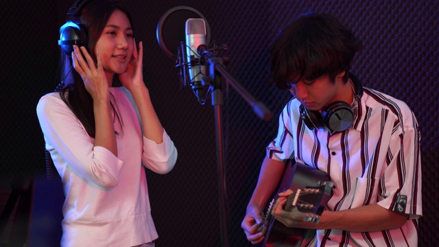 年轻的亚洲女人唱歌，而男人在背景弹吉他。年轻的当代音响操作员英俊的男性处理音乐。录音室混音歌曲乐队概念。视频素材