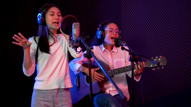 年轻的亚洲女人唱歌，而男人在背景弹吉他。年轻的当代音响操作员英俊的男性处理音乐。录音室混音歌曲乐队概念视频素材